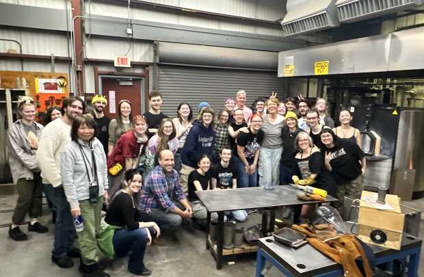 A group of about 30 smiling people pose together in an industrial MassArt workshop, surrounded by metal equipment and work tables. Some are standing, some kneeling, and many flash peace signs—a perfect stroll down memory lane.