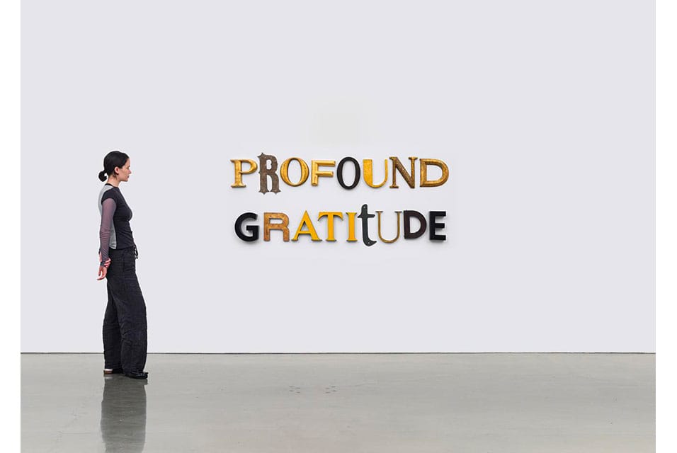 A woman stands in Regen Projects, looking at a Jack Pierson wall installation that spells out PROFOUND GRATITUDE in large, varied fonts and colors. The gallery features plain white walls and a polished concrete floor—part of his survey exhibition.
