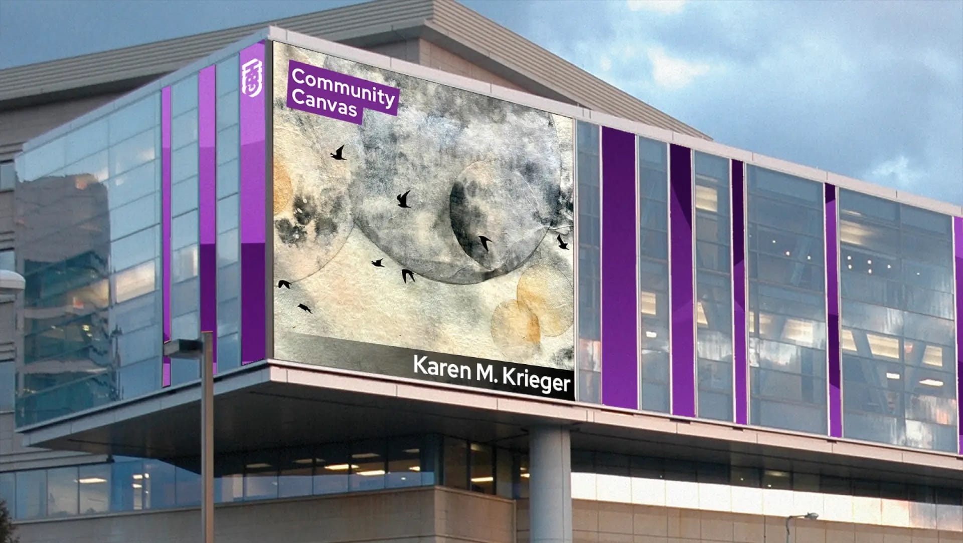 A modern building at MassArt displays a large outdoor mural titled Community Canvas by alum Karen M. Krieger, featuring abstract watercolor art with black birds and circular shapes. Purple accents frame the artwork on the building.