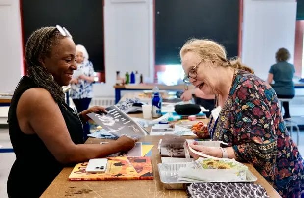 Two women smile and talk at a classroom table during Maker Days, with others working in the background.