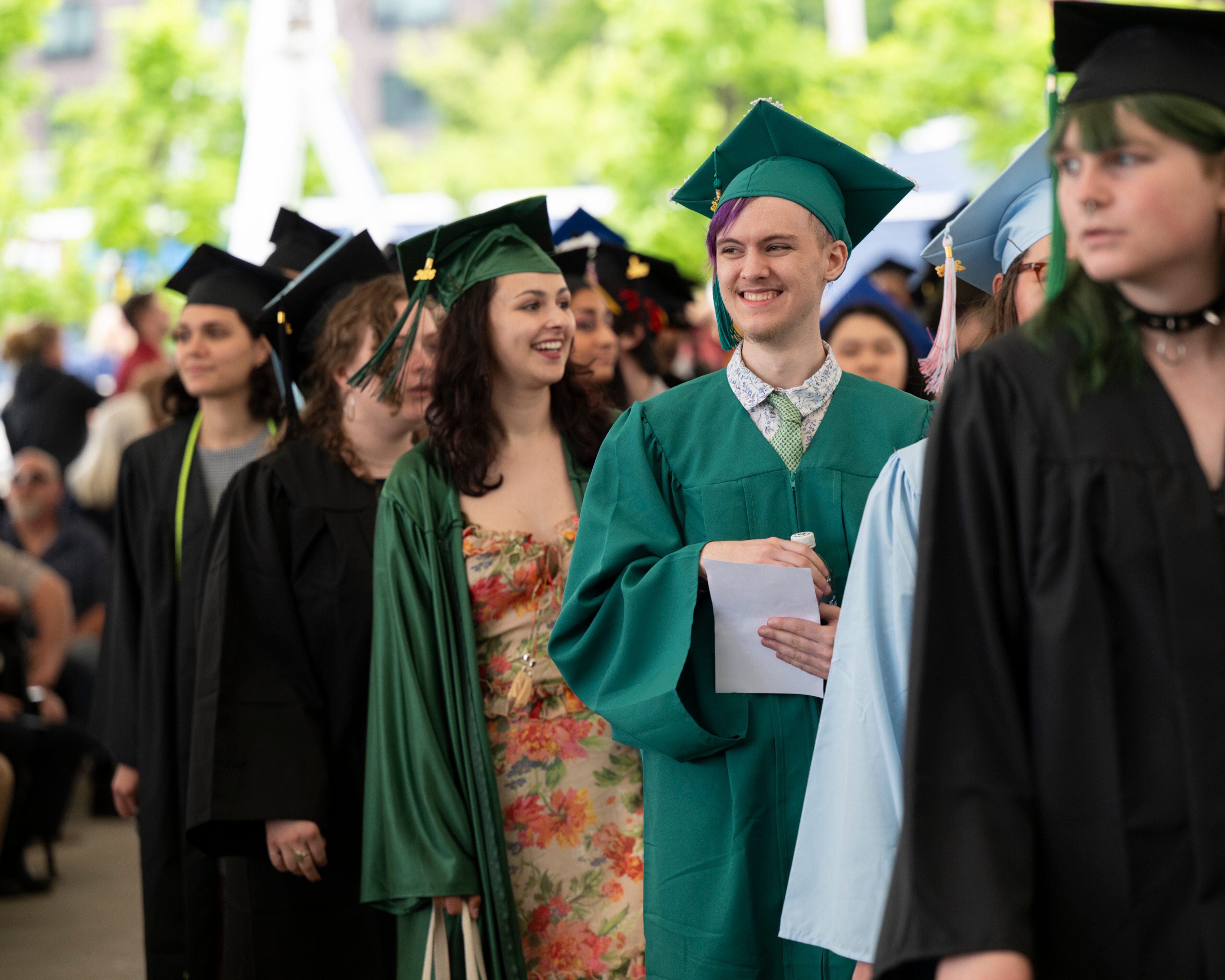 Graduates in gowns and caps stand in a line, smiling, at the MassArt Commencement ceremony outdoors.