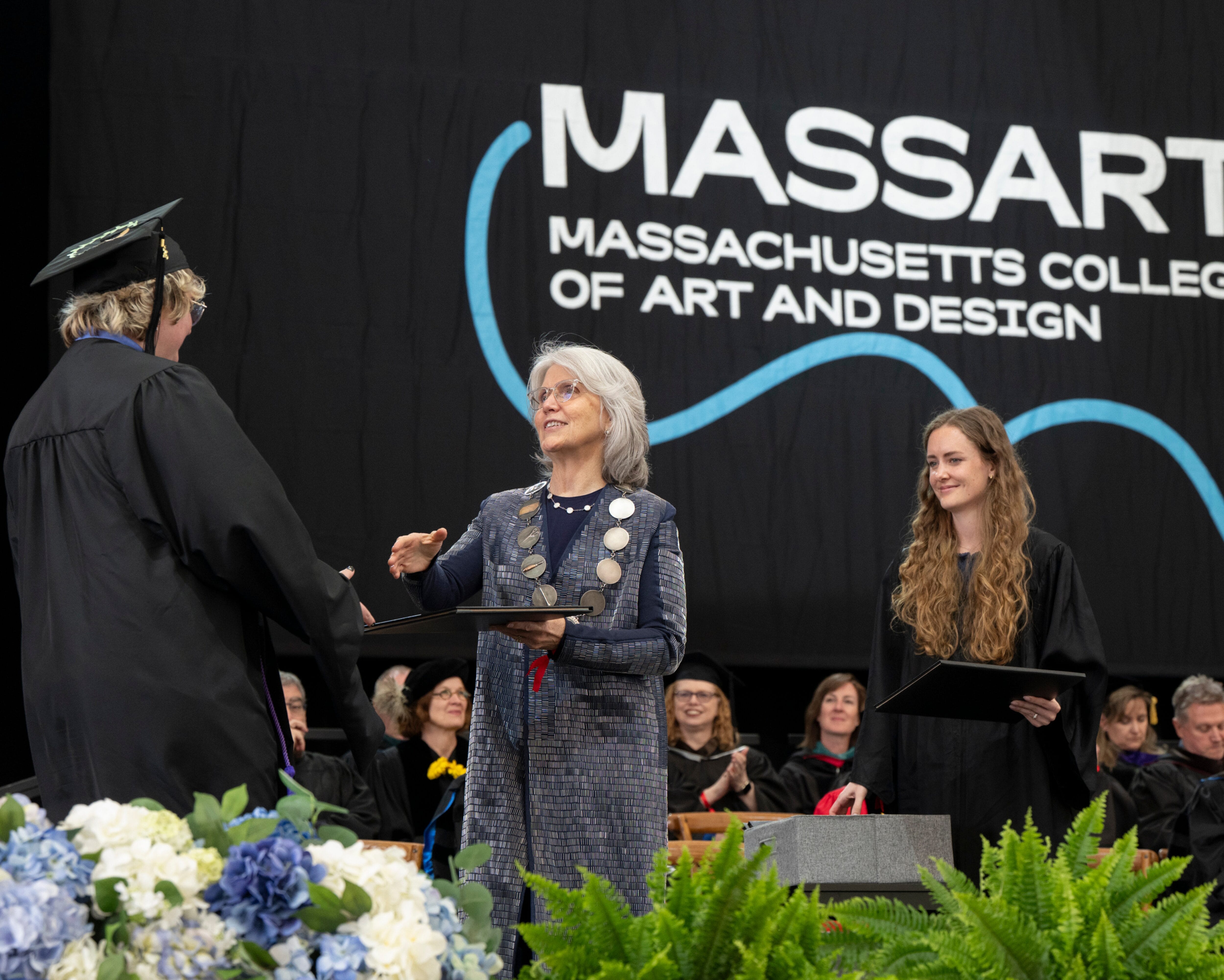 A graduate receives a diploma on stage during the MassArt Commencement ceremony, with officials and faculty present.