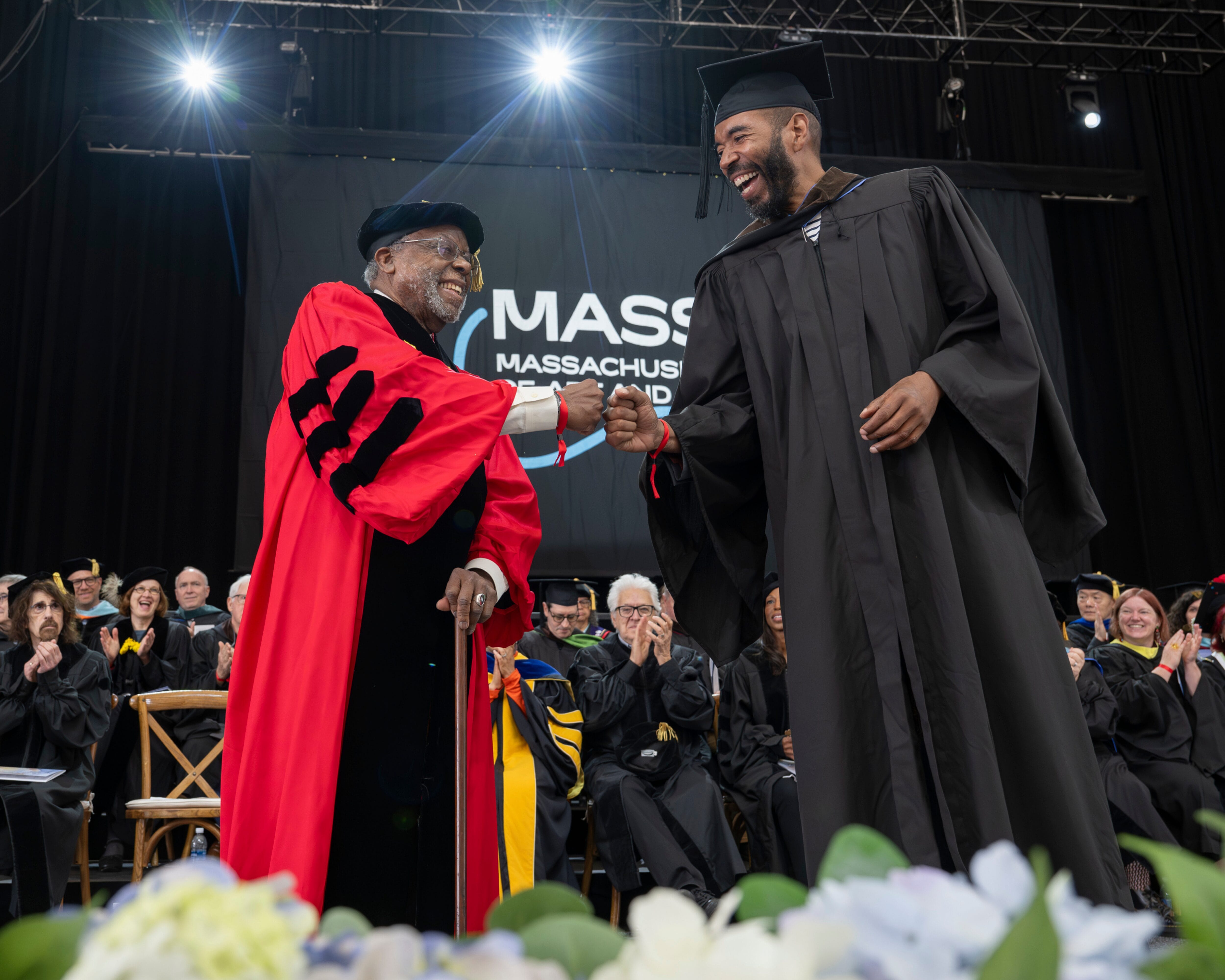 A graduate in cap and gown fist-bumps a faculty member in red robes on stage at MassArt Commencement.