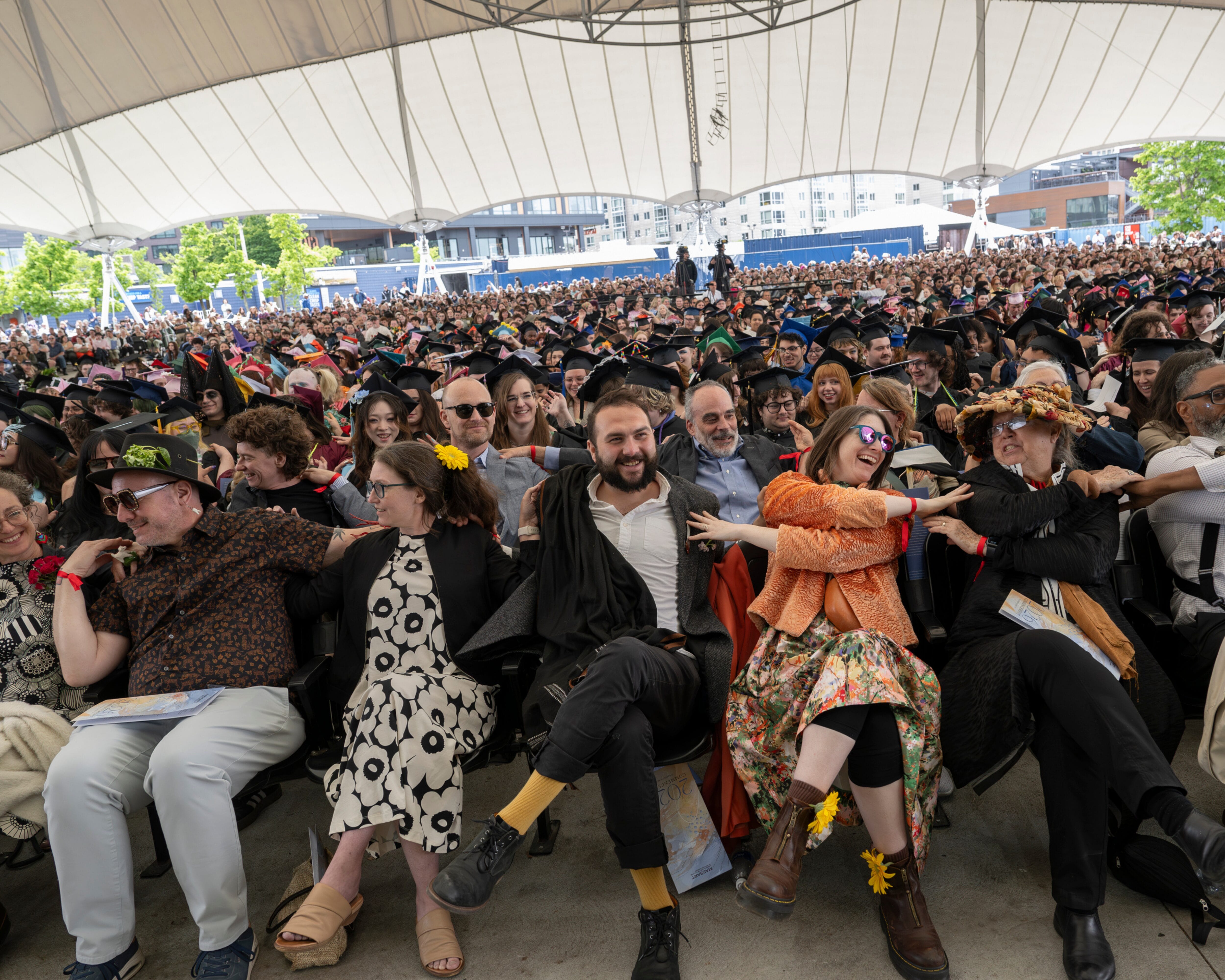 A large crowd in colorful outfits links arms at the outdoor MassArt Commencement under a canopy.