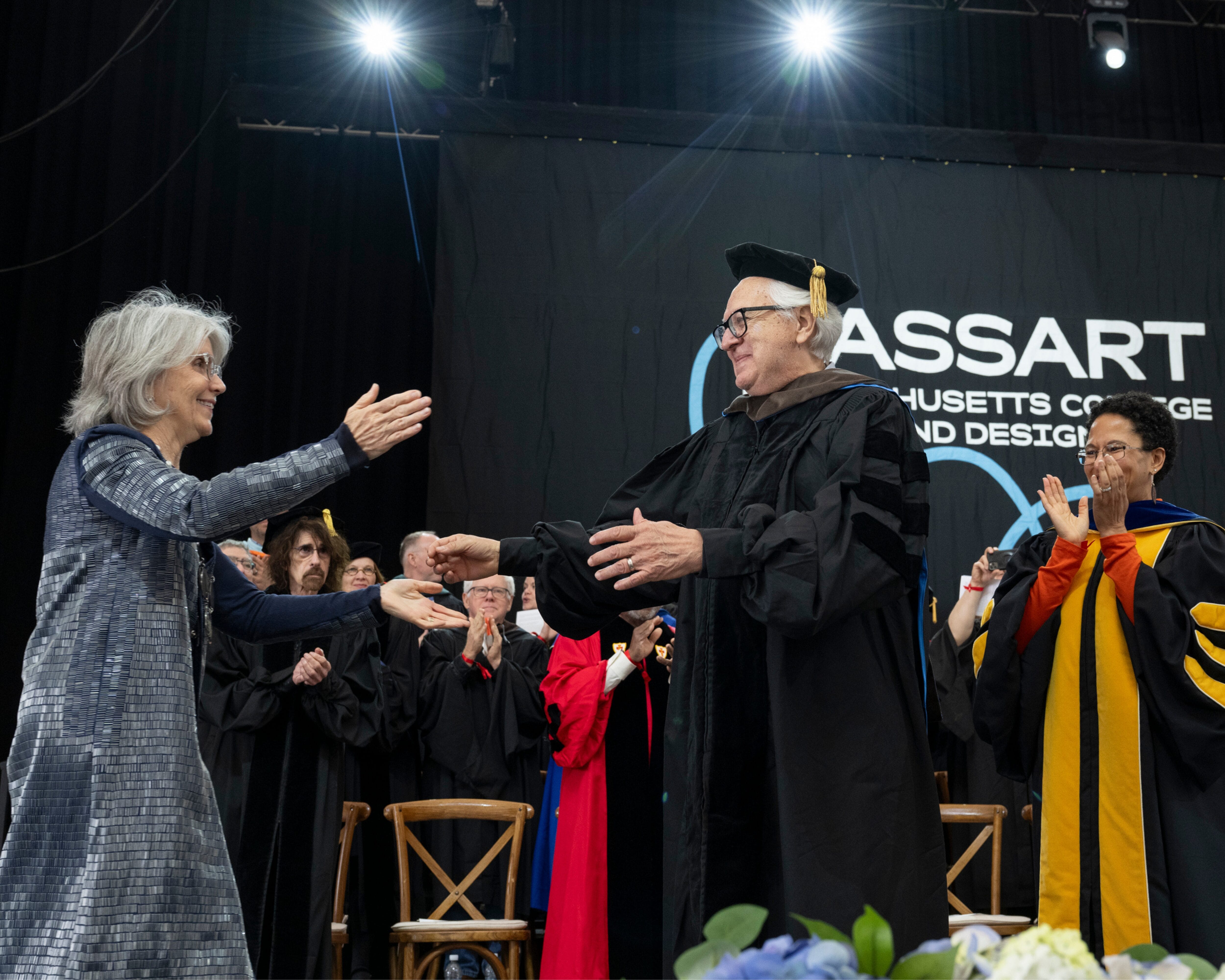 Two people in academic regalia shake hands on stage at MassArt Commencement as others clap in the background.