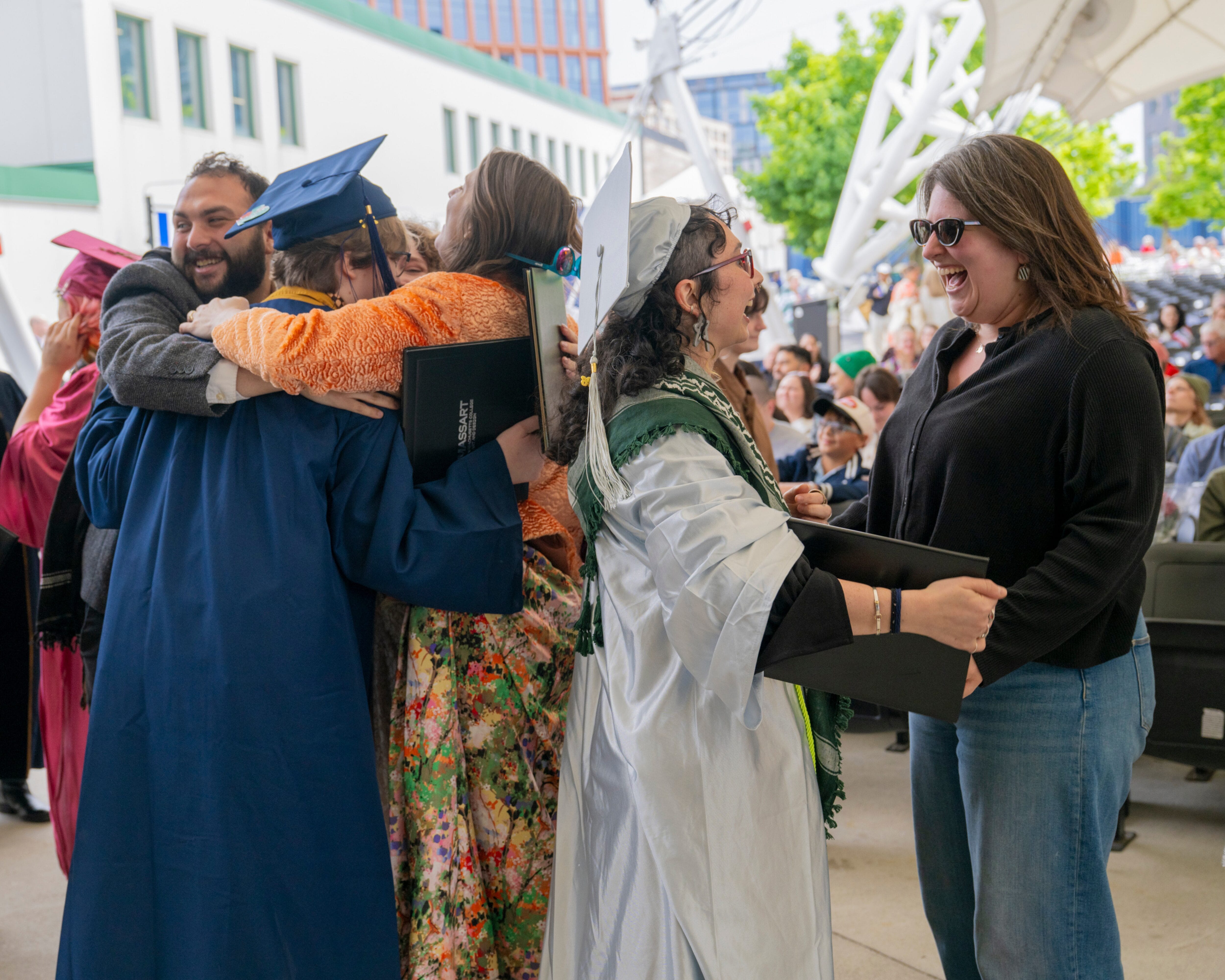 Graduates in caps and gowns hug and celebrate with loved ones at the MassArt Commencement ceremony outdoors.