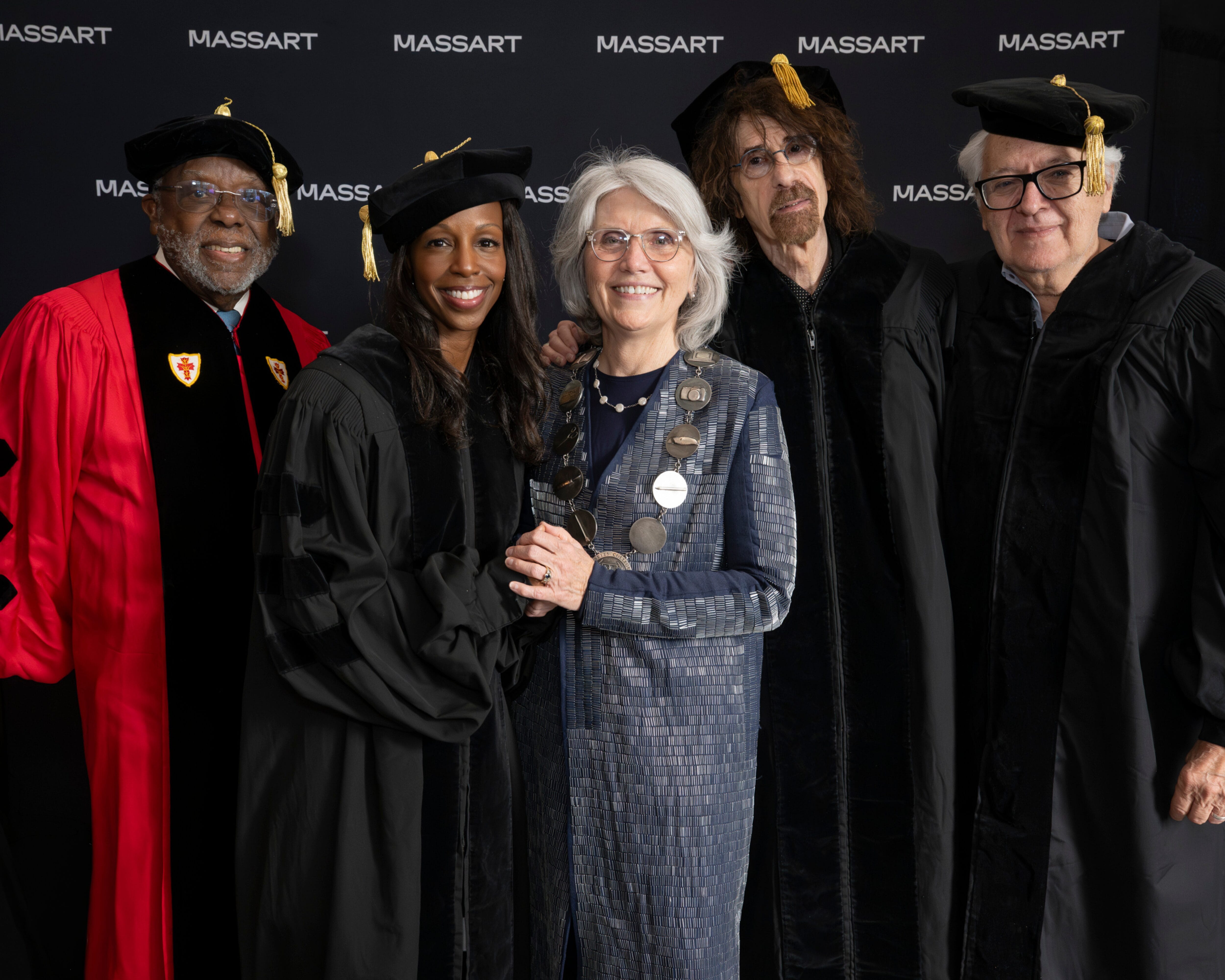 Five people in academic regalia smile together in front of a MassArt Commencement backdrop.
