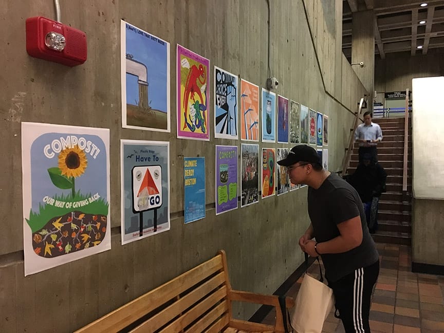 A person views colorful posters about sustainability, composting, and climate change on a concrete hallway wall indoors.
