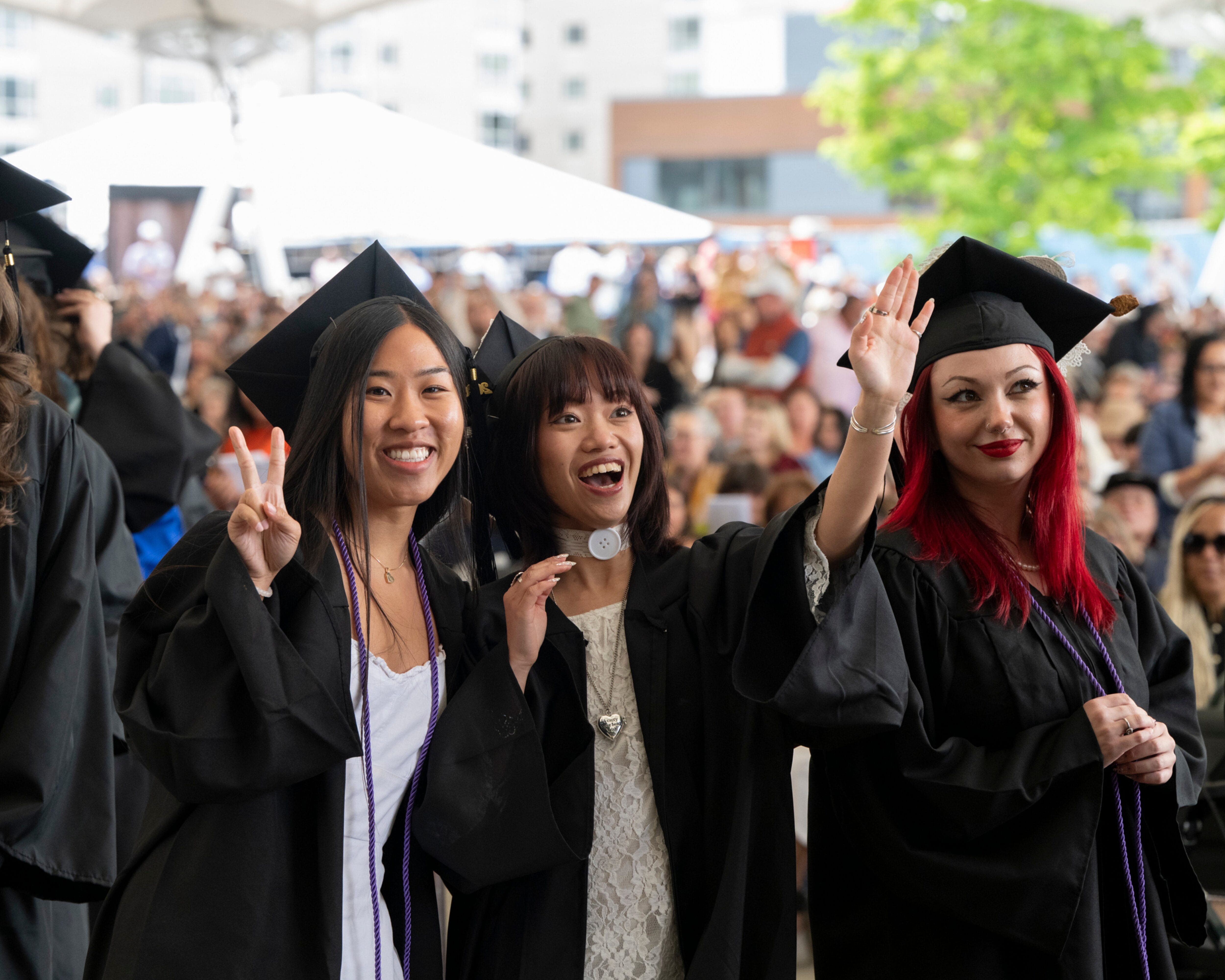 Three graduates in caps and gowns smile and wave at the camera during the MassArt Commencement ceremony outdoors.