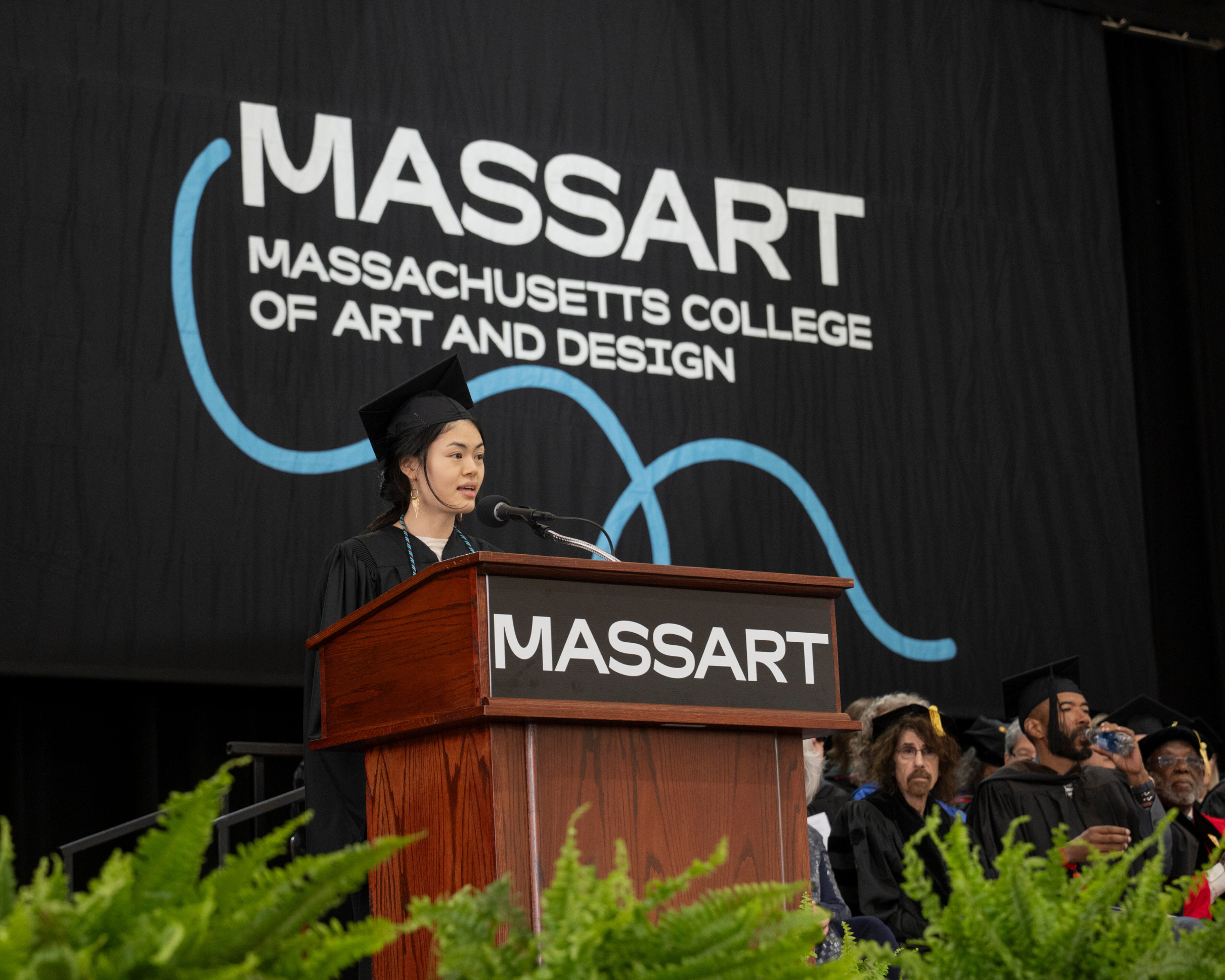 A graduate speaks at a podium during the MassArt Commencement ceremony, with others seated behind her on stage.