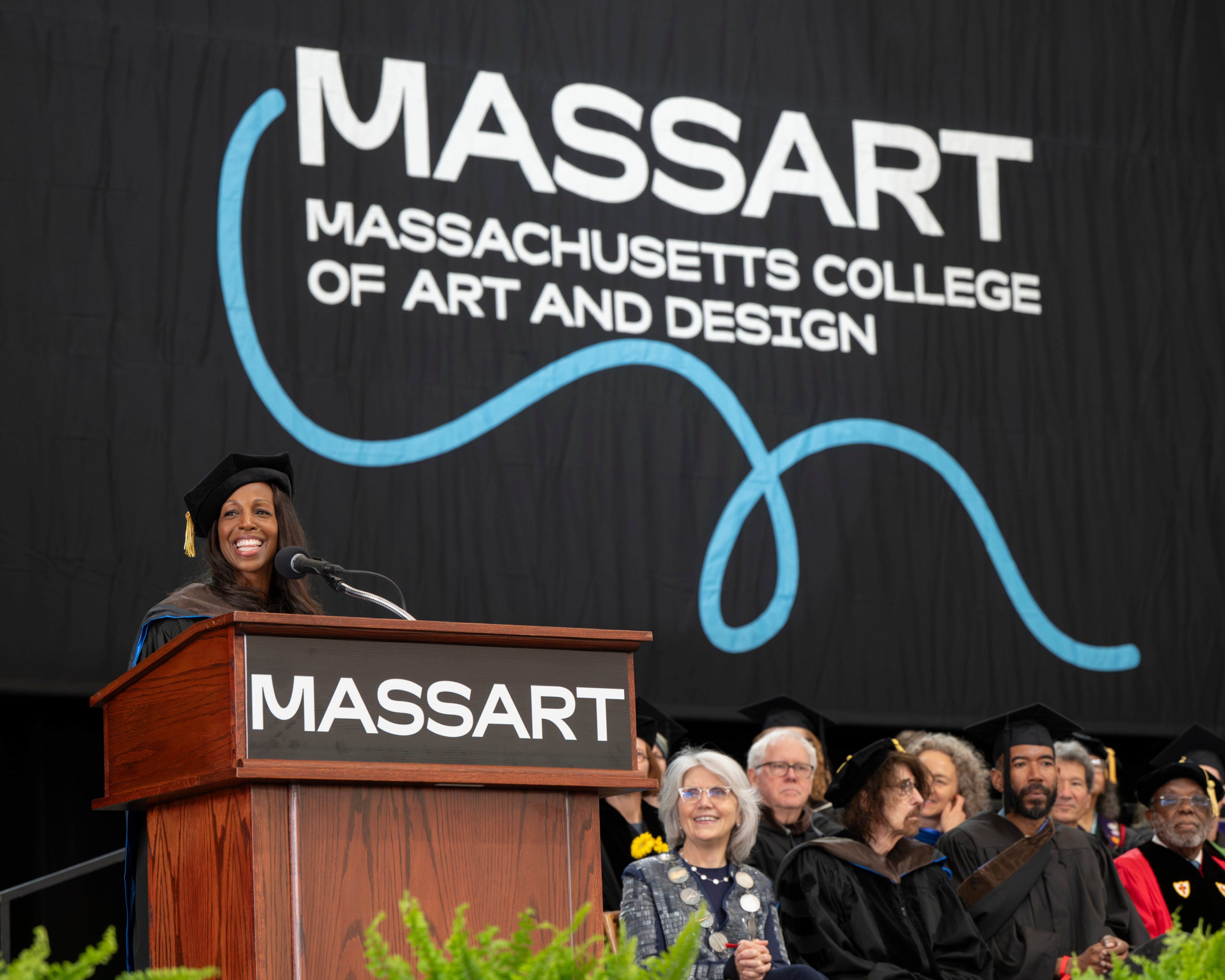 A speaker at a podium addresses the MassArt Commencement ceremony at Massachusetts College of Art and Design.