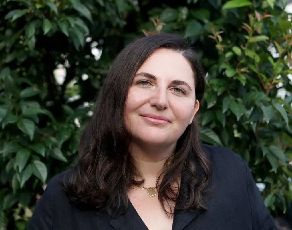 Woman with dark hair and a black top smiles softly while standing in front of leafy green bushes outdoors.