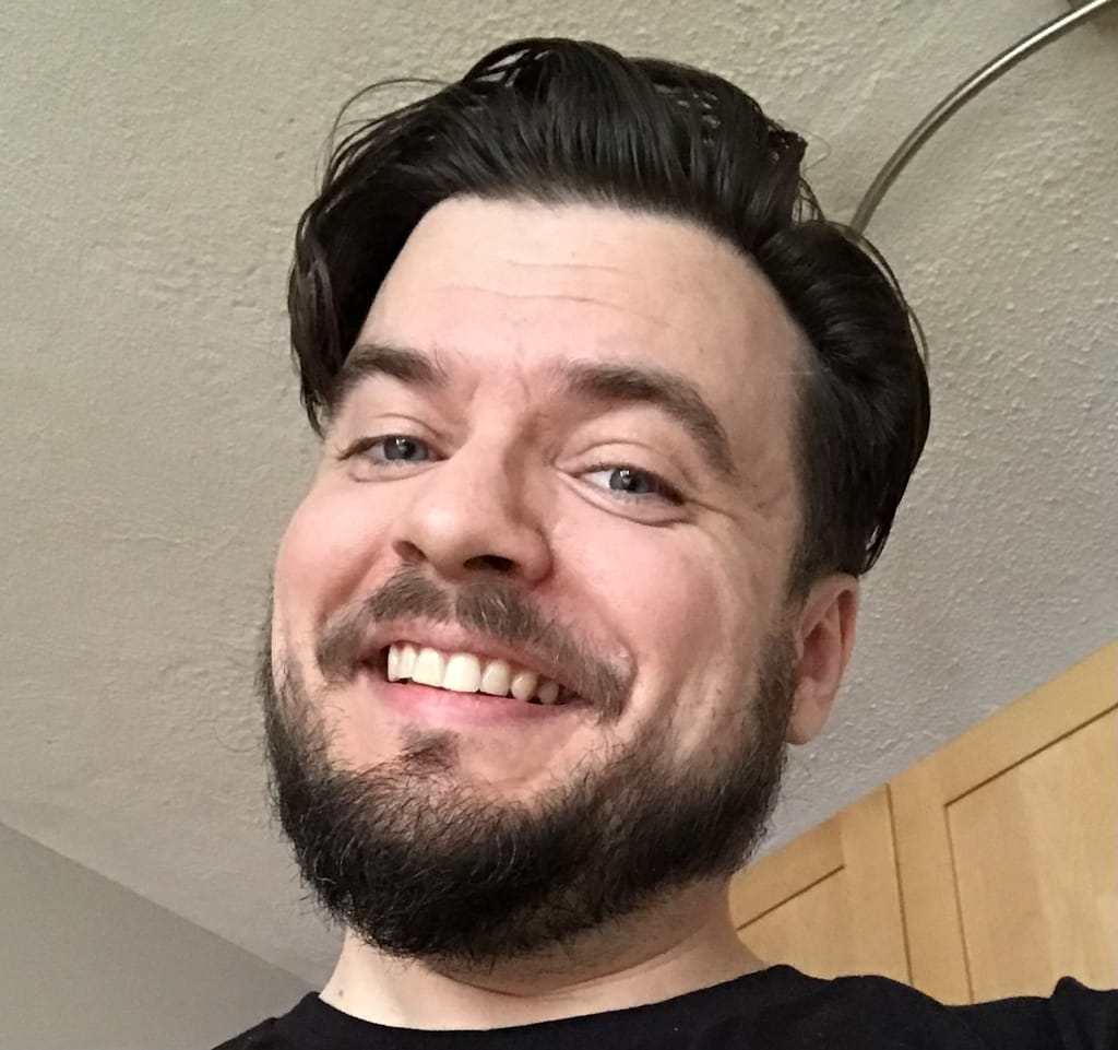 Smiling man with dark hair and beard, wearing a black shirt, standing indoors near a light-colored cabinet.