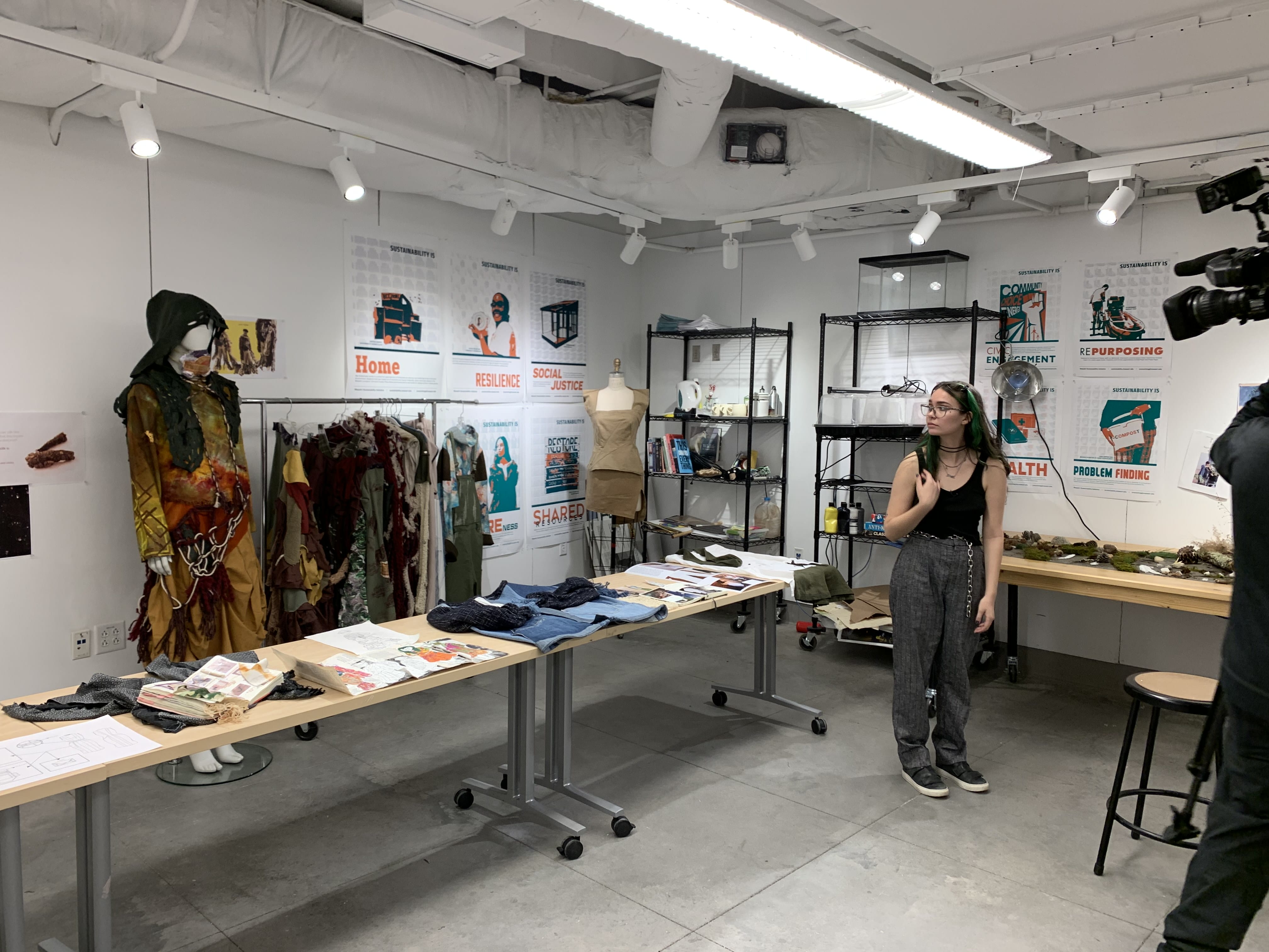 A woman stands in a well-lit MassArt exhibit with clothing, papers, and posters displayed around the Climate Beacon.