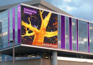 A large digital display on a modern building shows Jackie Powell’s Community Canvas illustration featuring yellow tree branches.