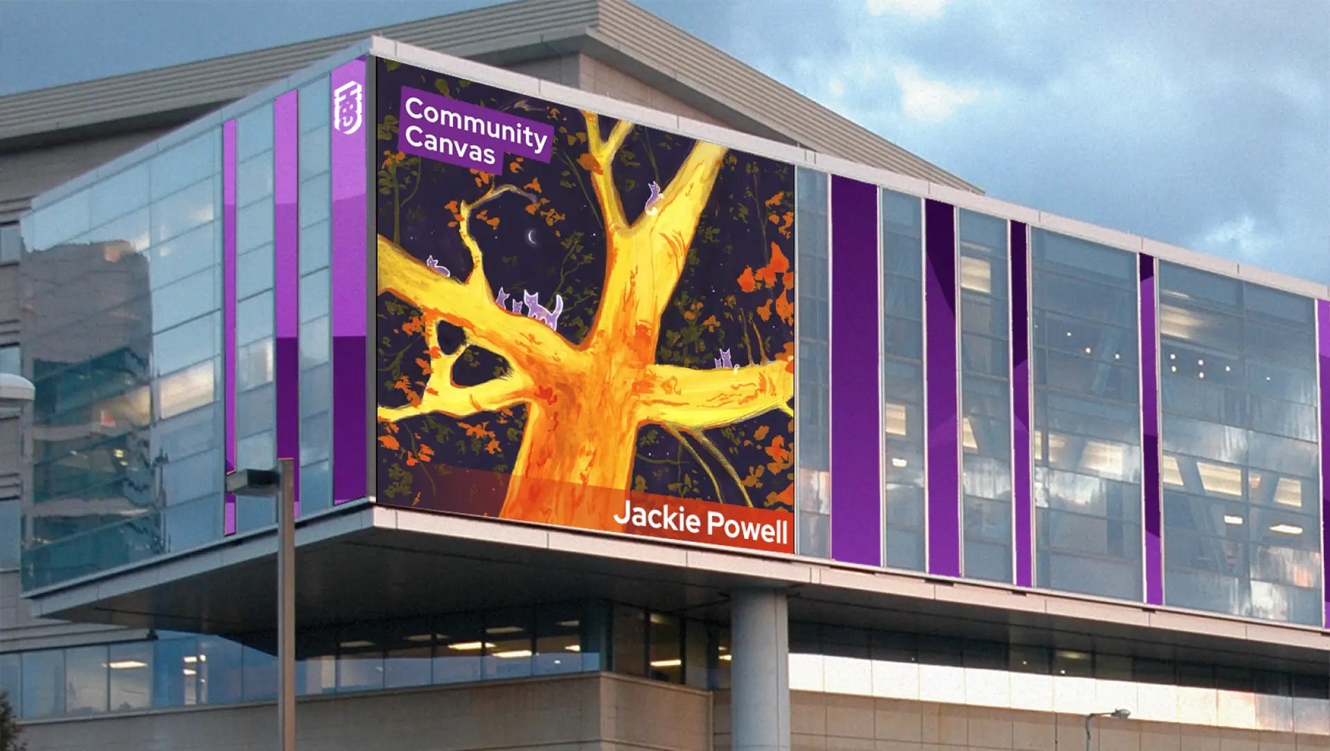 A large digital display on a modern building shows Jackie Powell’s Community Canvas illustration featuring yellow tree branches.