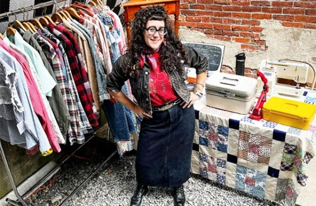 A smiling woman in vintage clothes stands by a rack of shirts and a table with sewing supplies outdoors by a brick wall.