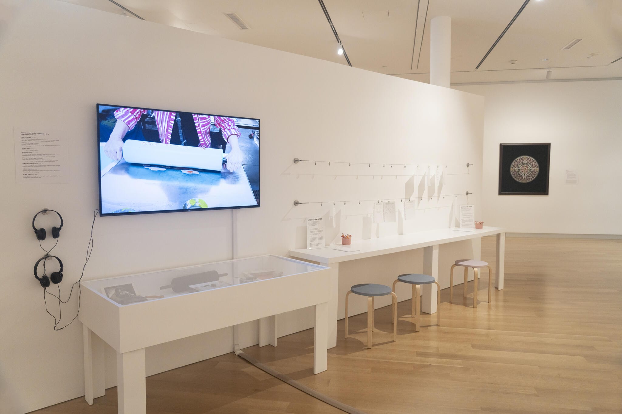 A modern art gallery at MassArt Art Museum with stools, TV screen, headphones, and a display table on wooden floors.